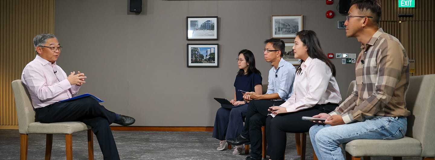 Six adults seated in a row, listening to a speaker on the left with a blue folder.